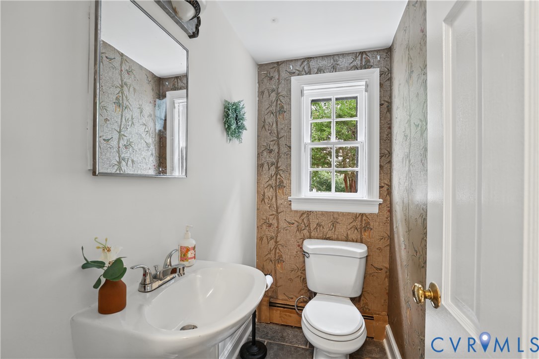 Powder room featuring patterned wallpaper, a wall-mounted sink with a chrome faucet, a rectangular framed mirror, a multi-pane window, and a standard-height toilet