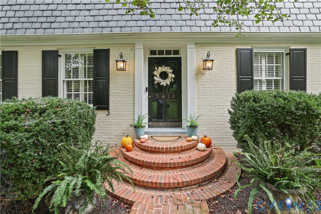 Brick exterior with shuttered windows, slate-style roofing, and curved masonry front steps leading to a central glass-paneled door flanked by wall-mounted lanterns