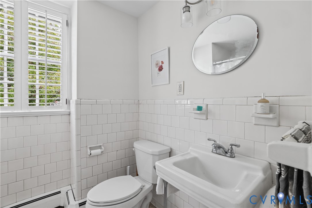 Wall-mounted porcelain sink with dual-handle metal faucet, oval wall mirror, white subway tile wainscoting, and window with built-in shutters