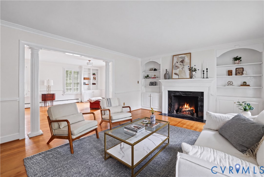 Living area featuring wood-finish flooring, a fireplace with a dark stone surround and white mantel, symmetrical built-in shelving, and architectural columns