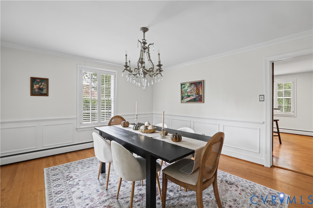Dining area featuring wood-finish flooring, white wainscoting, multi-pane windows with plantation shutters, and a tiered crystal chandelier