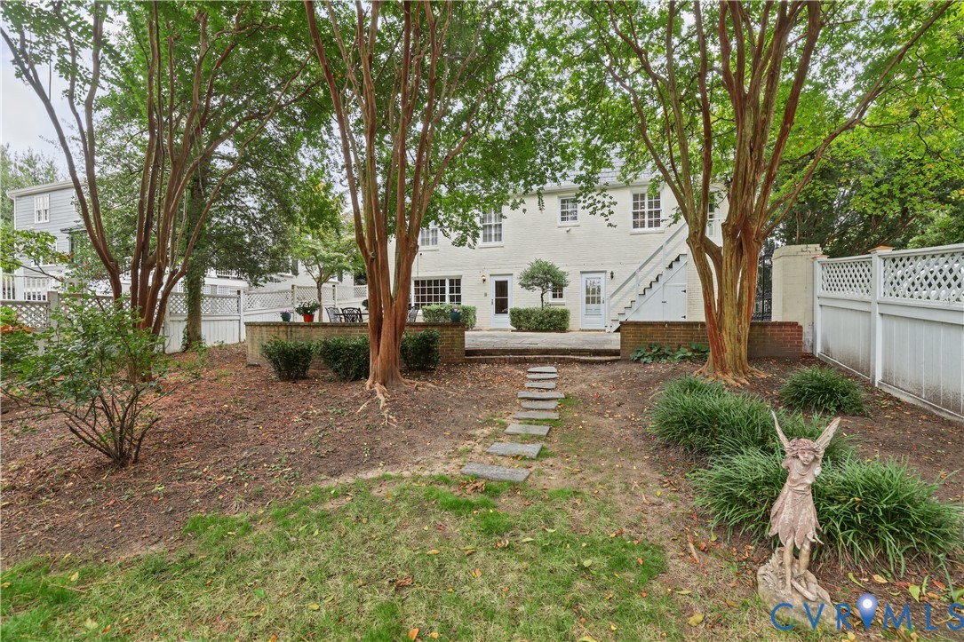 Backyard featuring a paved stone patio, retaining wall, stone paver pathway, mature trees, and white lattice fencing