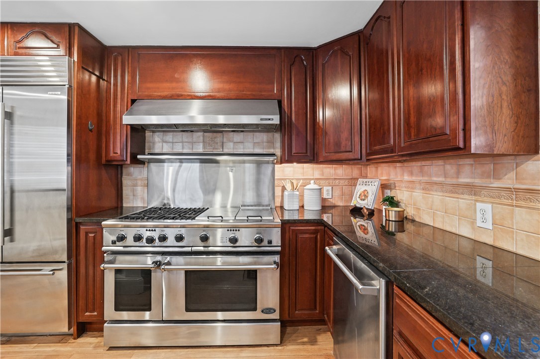 Kitchen featuring custom cherry-toned cabinetry, stainless steel professional-grade gas range, matching stainless steel hood, built-in refrigerator, and stone countertops with a ceramic tile backsplash