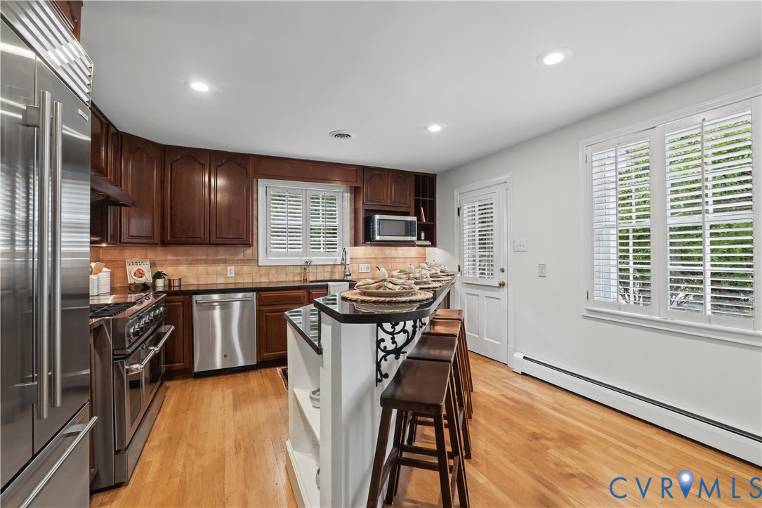 Kitchen featuring dark wood cabinetry, black countertops, stainless steel appliances, wood-finish flooring, and plantation shutters