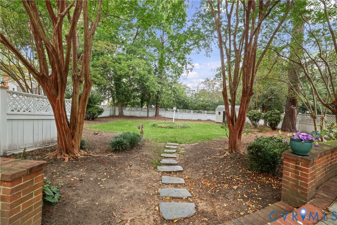 Spacious backyard featuring mature trees, a central lawn area, stone pathway, brick retaining wall, and white lattice fencing