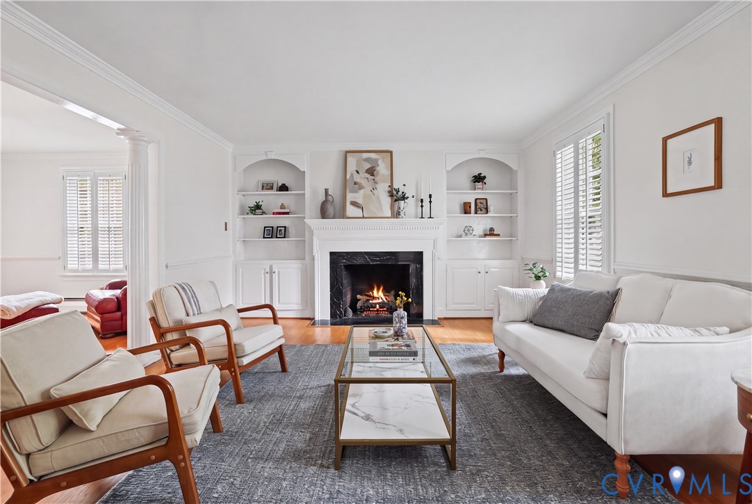 Living room featuring polished wood-finish flooring, built-in recessed cabinetry, crown molding, plantation shutters, and a fireplace with a stone surround