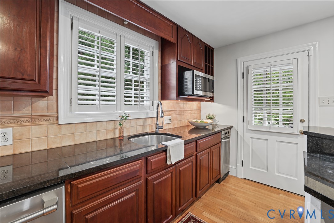 Stained wood cabinetry, stone countertops, tiled backsplash, stainless steel sink with gooseneck faucet, and wood-finish flooring
