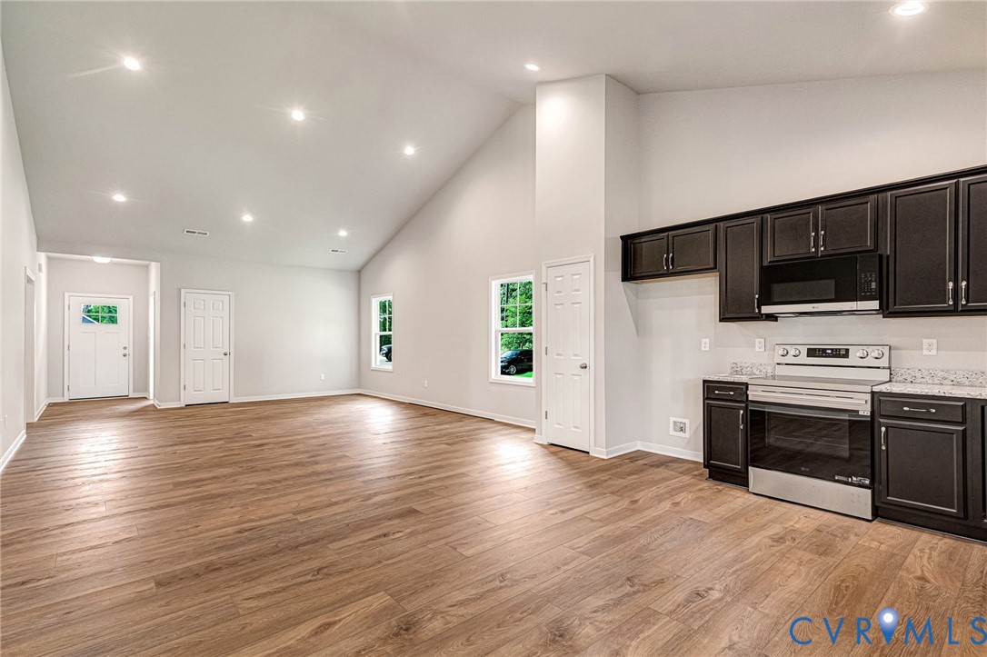 Open-concept living space with vaulted ceilings, recessed lighting, wood-finish flooring, and a kitchen featuring dark cabinetry, stainless steel appliances, and light-toned countertops