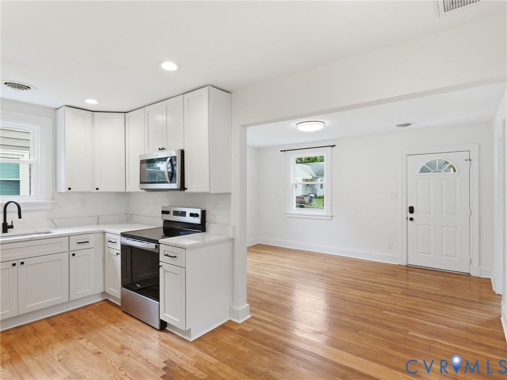 Bright kitchen and open living area featuring white cabinetry, stainless steel appliances, wood-finish flooring, recessed lighting, and a front entry door with an arched window insert