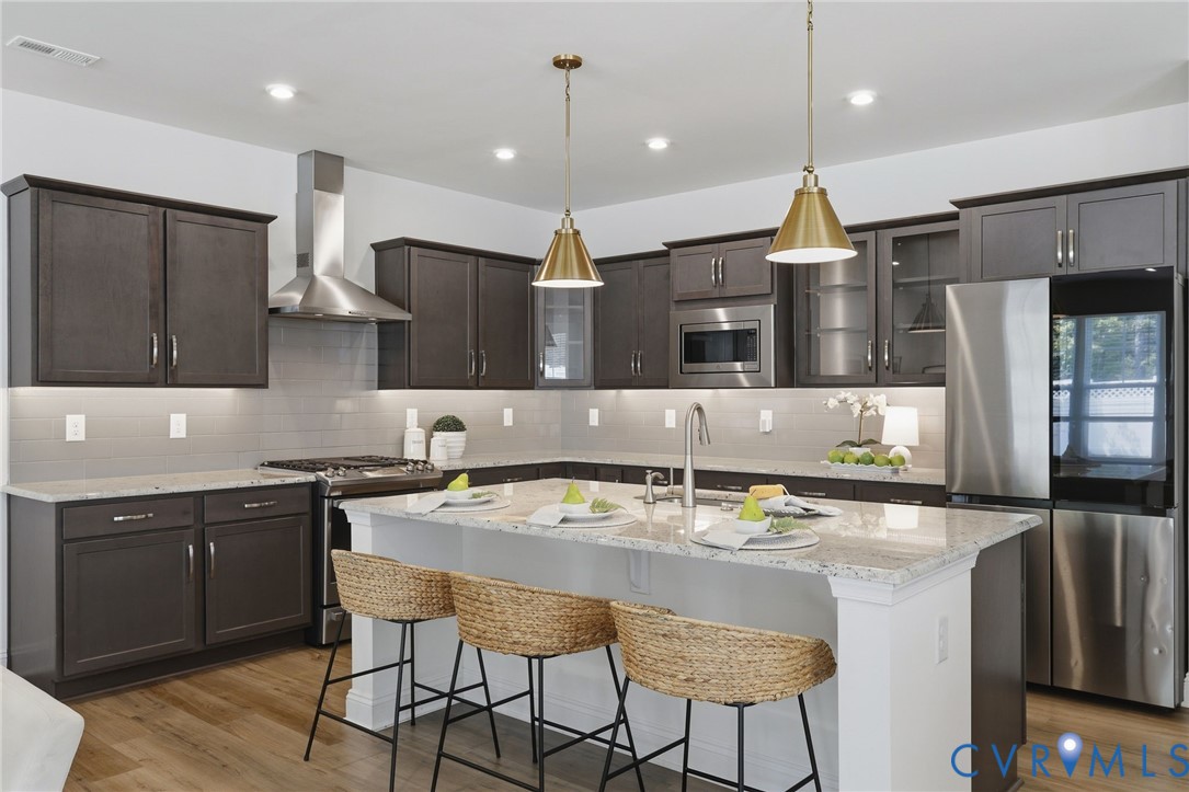 Spacious kitchen featuring dark wood cabinetry, stainless steel appliances, a large island with a light-toned countertop, a tile backsplash, and recessed and pendant lighting