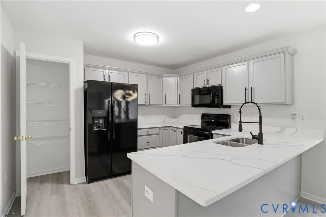 Modern kitchen featuring light gray cabinetry, black appliances, light wood-finish flooring, a white countertop island with an undermount sink, and a walk-in pantry with wire shelving