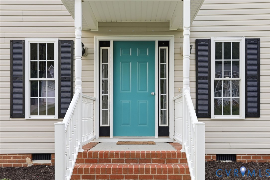 Welcoming front entry featuring a vibrant teal door, white vinyl siding, dark louvered shutters, brick steps, and white railings