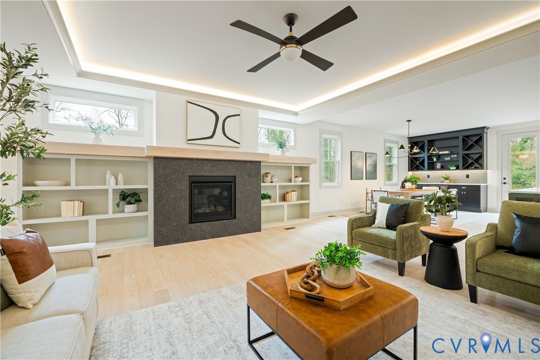 Spacious living area featuring a dark stone-clad fireplace with a natural wood mantel, built-in shelving, light wood-finish flooring, and a tray ceiling with recessed lighting Spacious living area featuring a dark stone-clad fireplace with a natural wood mantel, built-in shelving, light wood-finish flooring, and a tray ceiling with recessed lighting