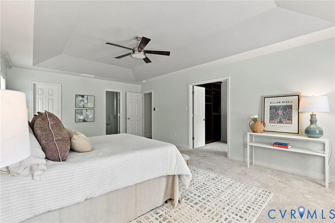 Tray ceiling with ceiling fan, light-colored wall paint, light-colored carpeting, and a doorway leading to a walk-in closet