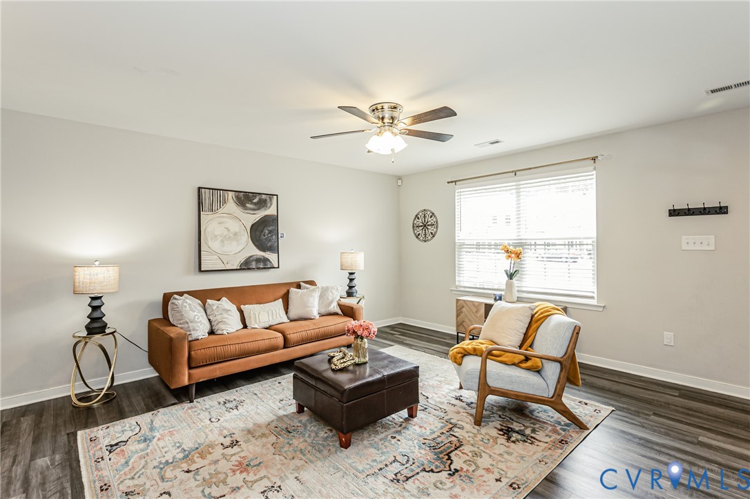 Spacious living area featuring wood-finish flooring, a ceiling fan with integrated lighting, and a large window with blinds