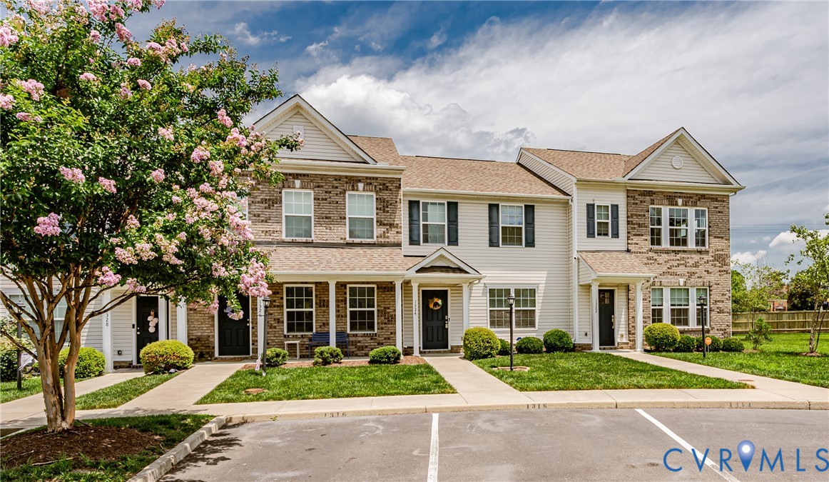 Two-story townhome exterior featuring light-colored siding, brick accents, dark shutters, and a covered porch entry