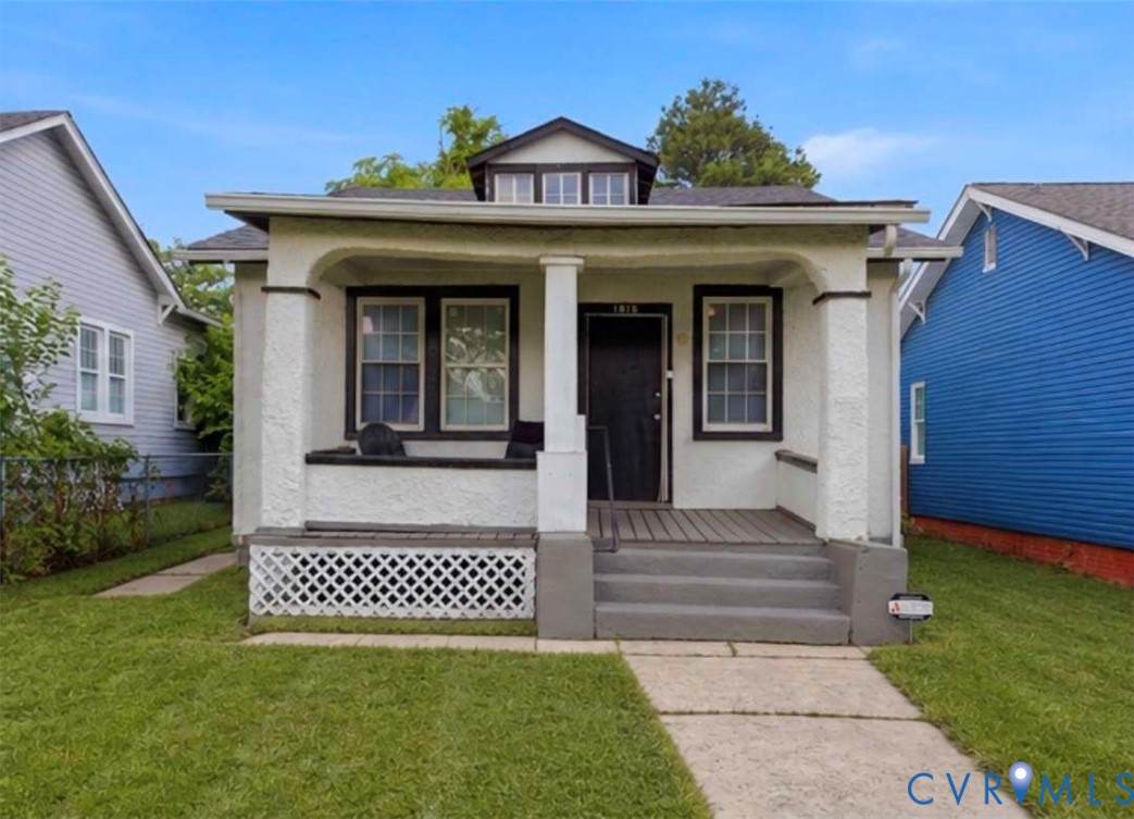 Front exterior featuring a covered porch with a dark wood-finish deck, stucco siding, a black front door, and a dormer window Front exterior featuring a covered porch with a dark wood-finish deck, stucco siding, a black front door, and a dormer window