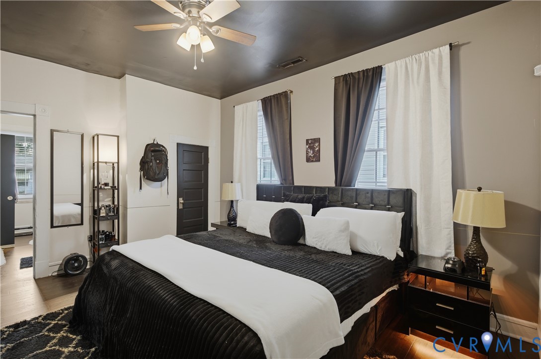 Bedroom featuring a ceiling fan with light kit, two windows with curtain rods, a paneled door, and wood-finish flooring