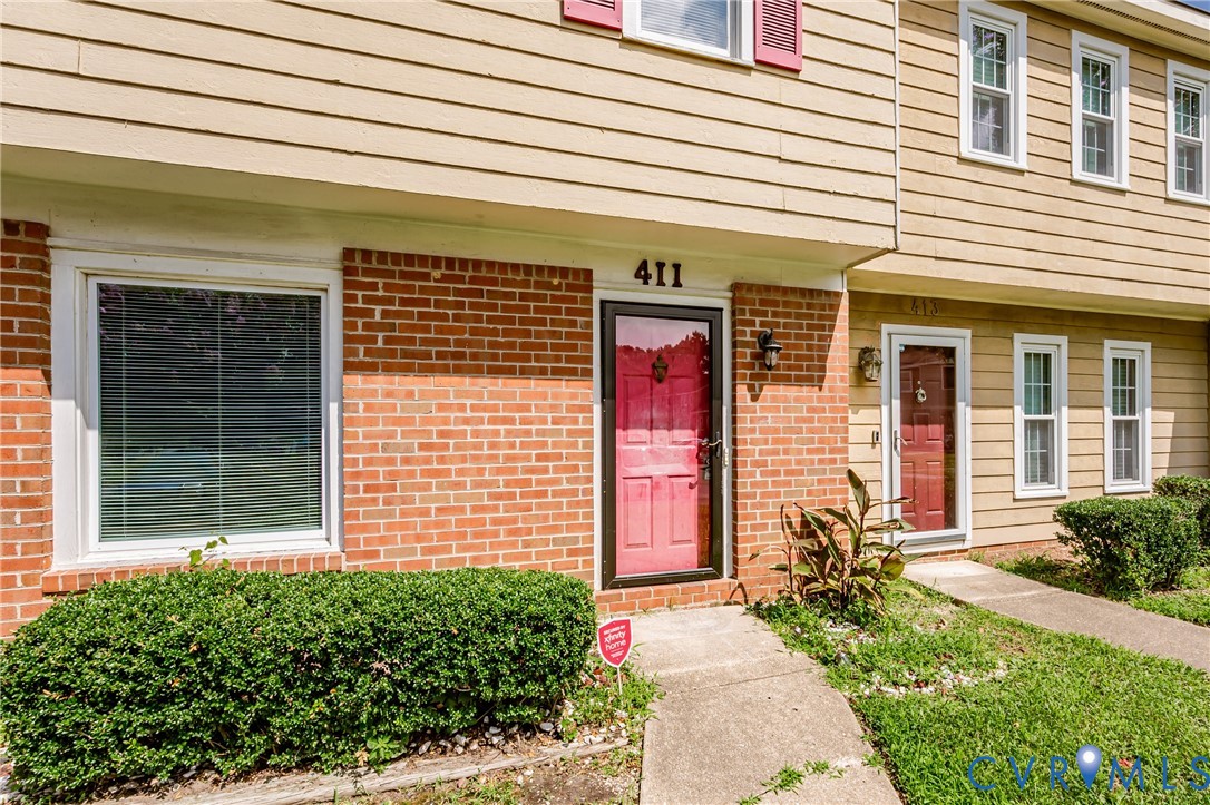 Brick and siding facade featuring a red entry door with a storm door, an exterior wall light, and a prominent front window with white blinds