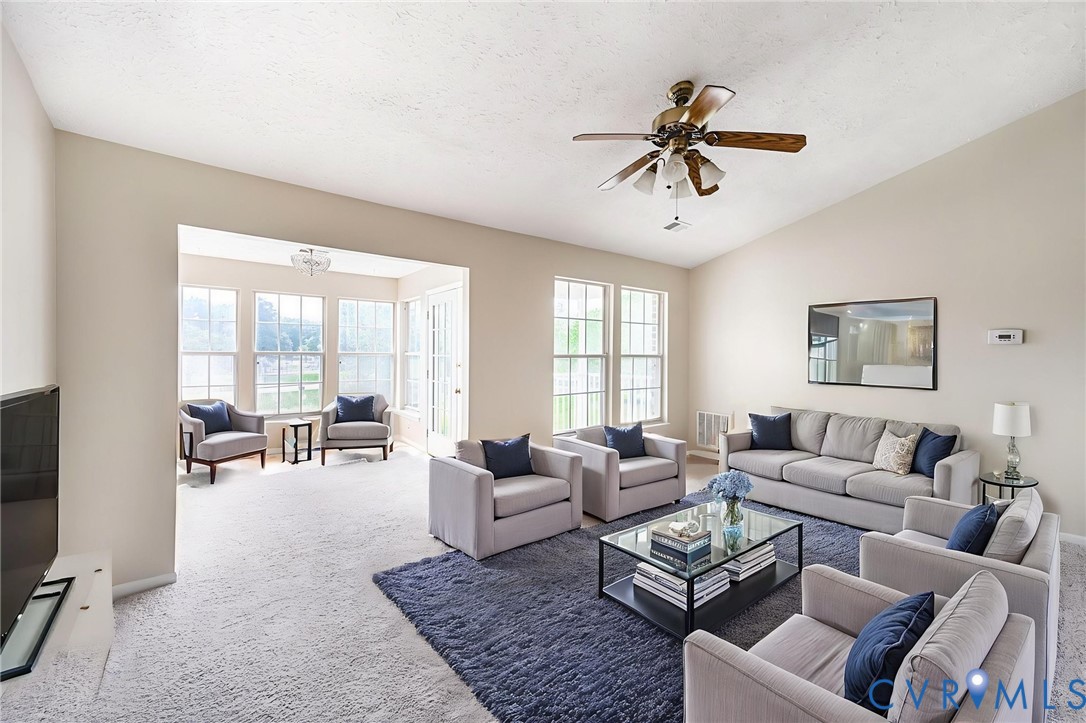 Spacious living room featuring a vaulted ceiling with a ceiling fan, light-colored carpeting, and multiple large windows~ Virtually Staged