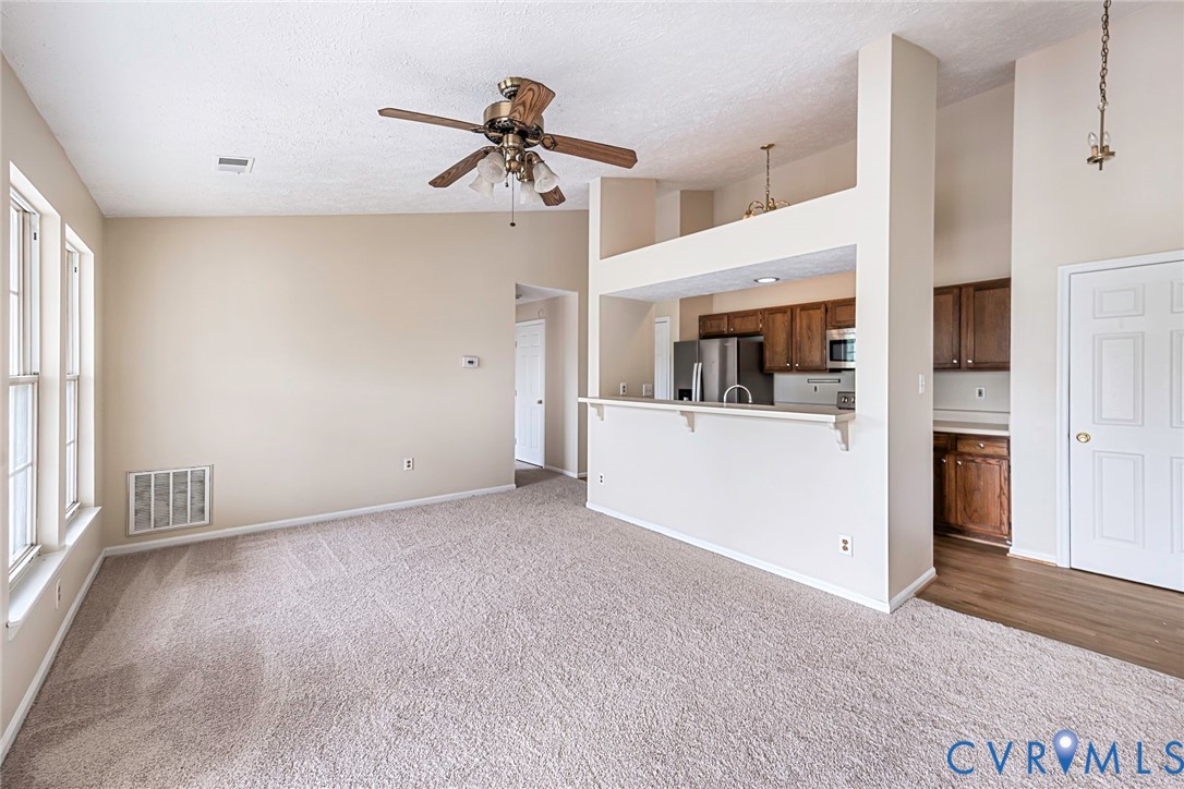 Expansive living area featuring light-toned carpet, a ceiling fan, and multiple windows
