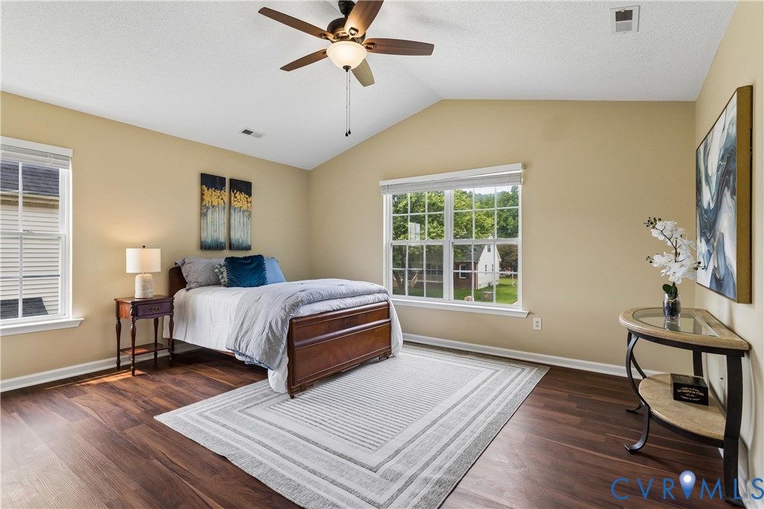 Vaulted ceiling bedroom featuring dark wood-finish flooring, a ceiling fan with light fixture, and multiple windows with blinds