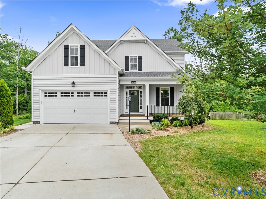 Two-story home featuring light-colored siding, a covered front porch with dark railings, a two-car garage with paneling and window accents, a concrete driveway, and landscaping with mature trees and shrubs