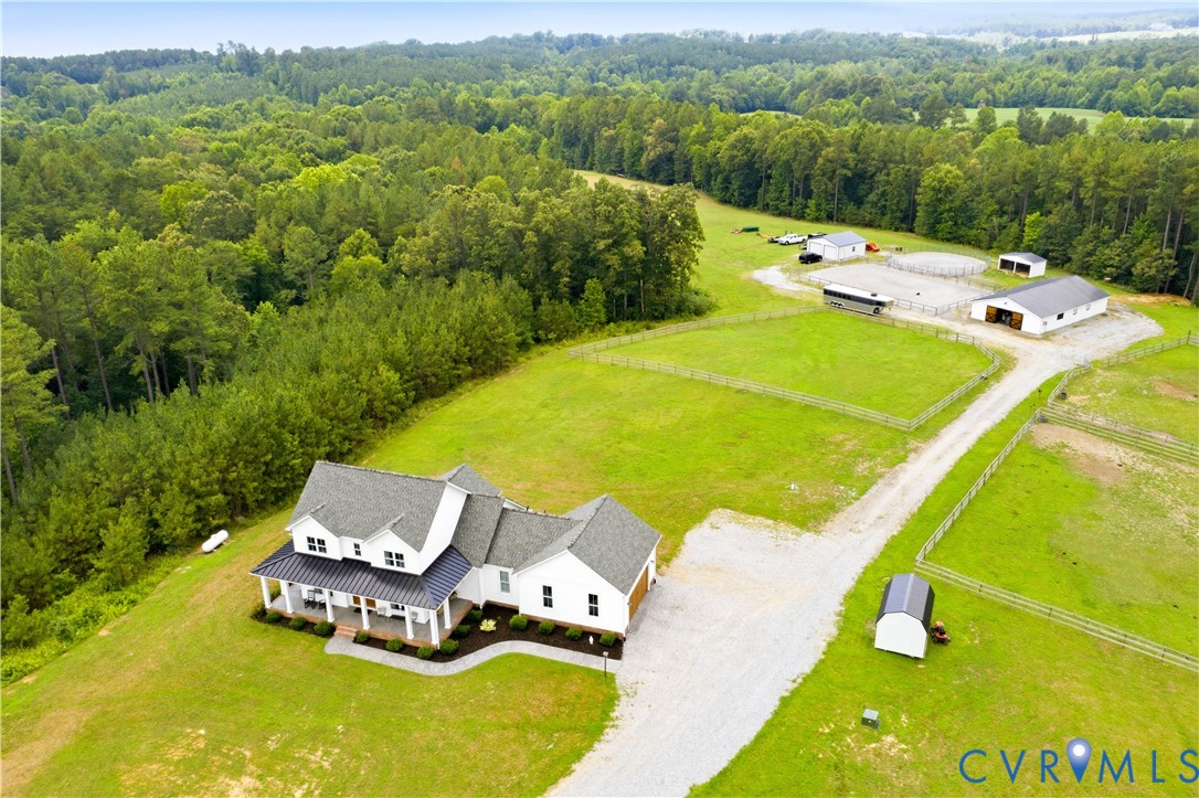 Expansive property featuring a white two-story residence with a covered front porch, gravel driveway, multiple outbuildings, fenced pastures, and a circular riding arena, all surrounded by mature woodlands
