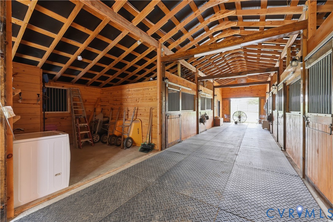 Wood-paneled barn interior featuring multiple stalls with metal grates, exposed beam ceiling, and durable rubber flooring