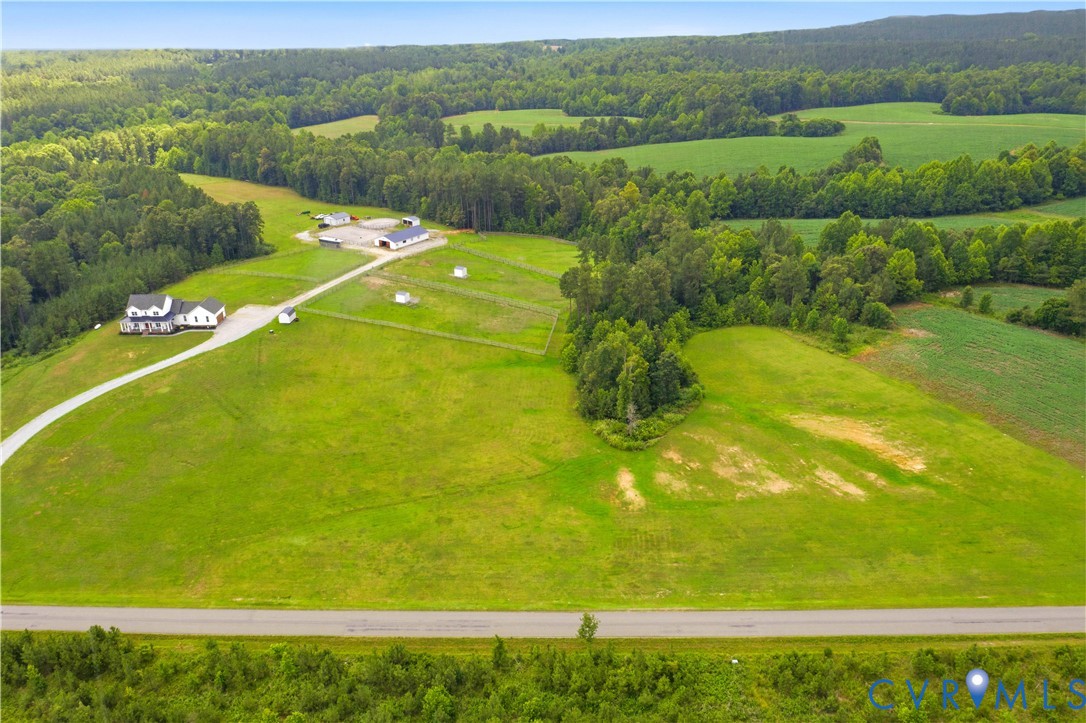 Expansive rural property featuring a white residence with a wrap-around porch, multiple outbuildings, a long gravel driveway, and extensive green fields bordered by mature trees