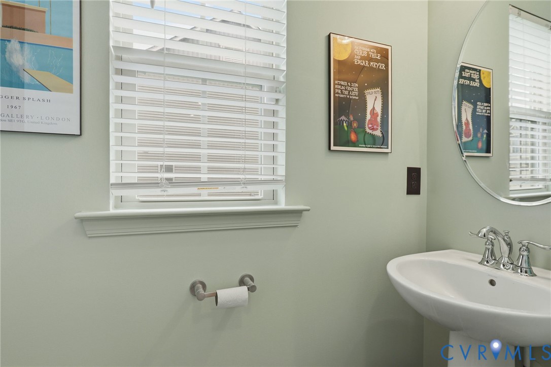 Pedestal sink featuring a chrome faucet, round mirror, and a window with blinds