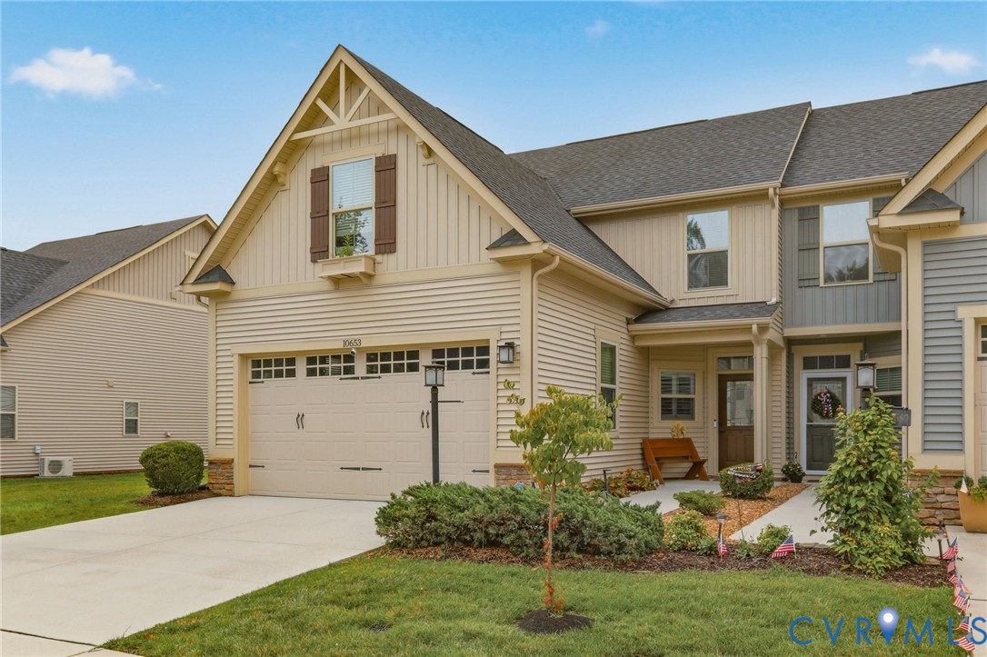 Townhome exterior featuring tan siding with stone accents, an attached garage, a covered front entry, and mature landscaping