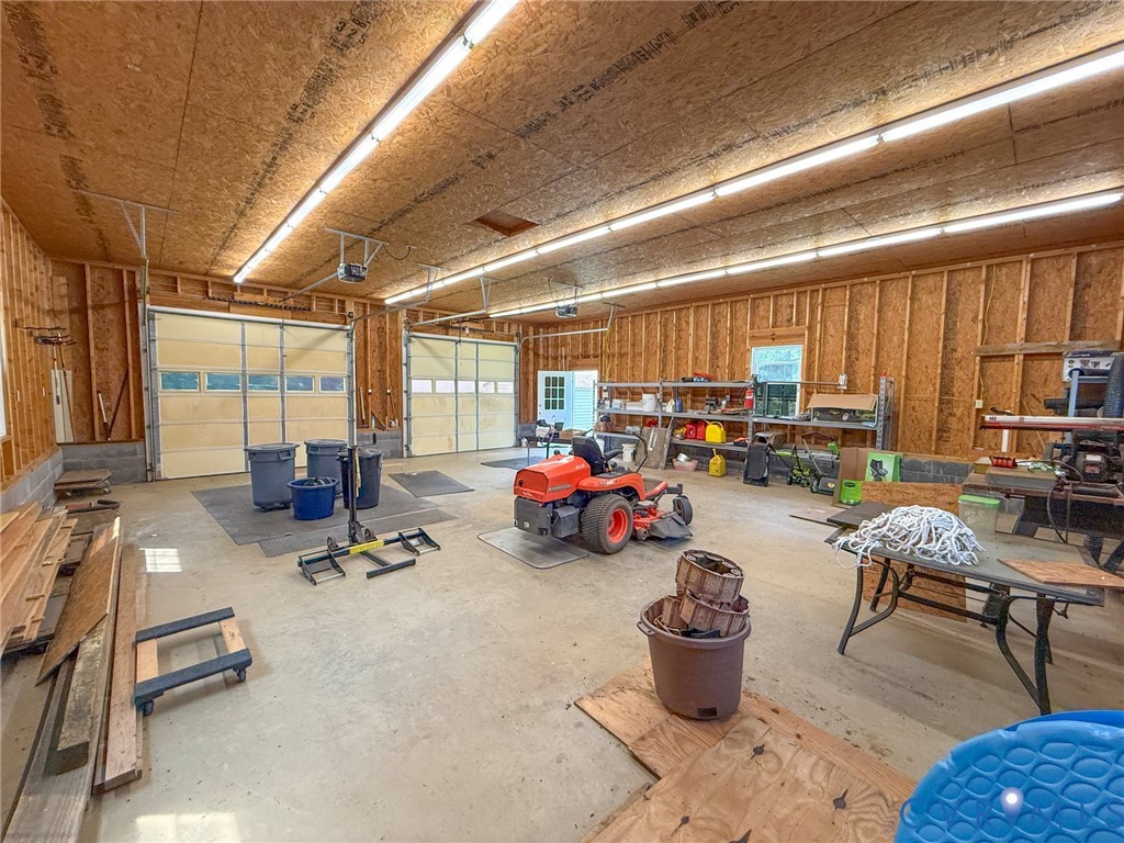 Spacious utility building featuring two roll-up garage doors, exposed wood-frame construction, concrete flooring, and extensive overhead strip lighting