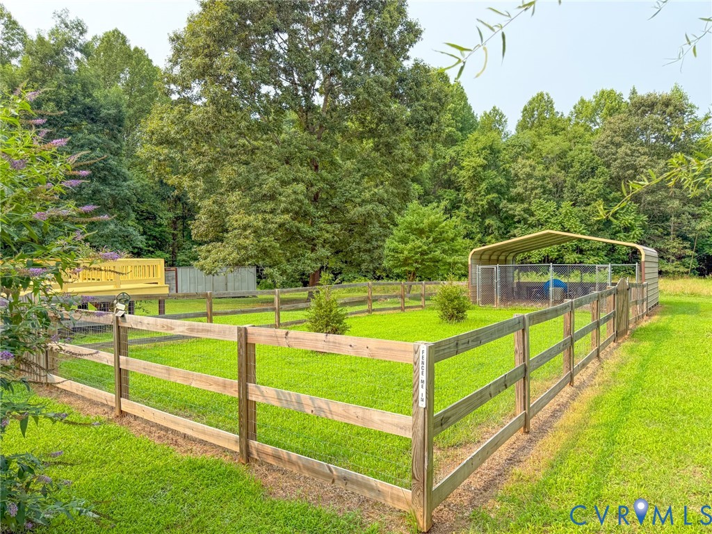 Expansive fenced lawn area featuring a wooden split-rail fence, a raised wood deck, a metal carport, and a separate fenced enclosure