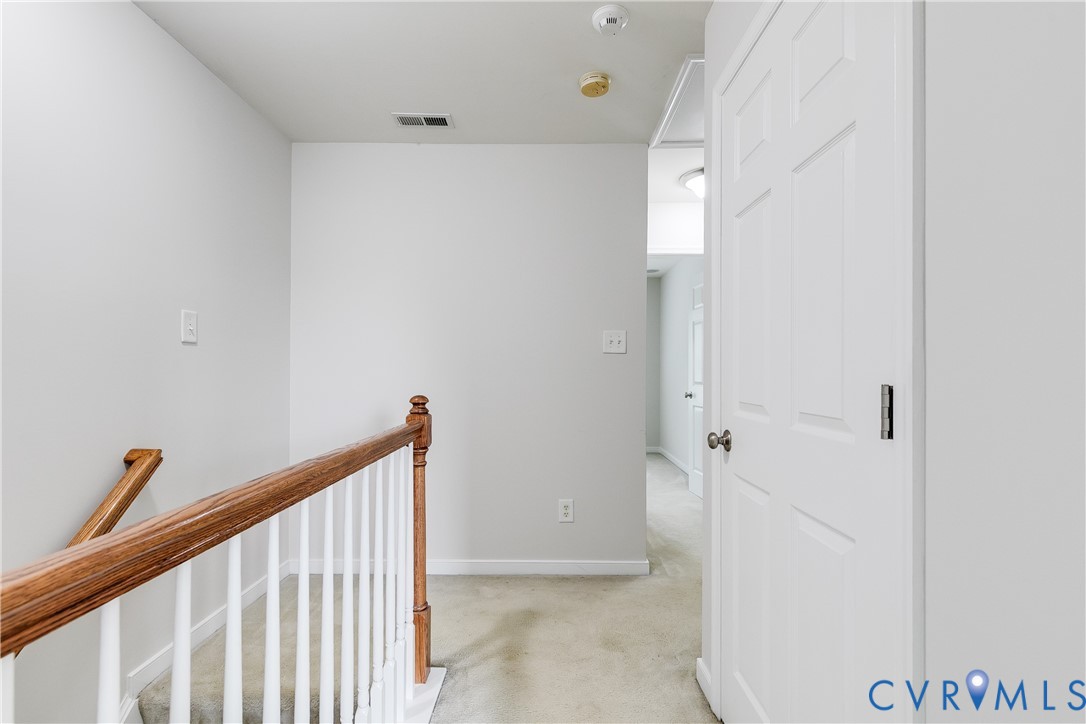 Upper-level hallway featuring light gray walls, carpet flooring, a white paneled door, and a wooden stair railing with white spindles