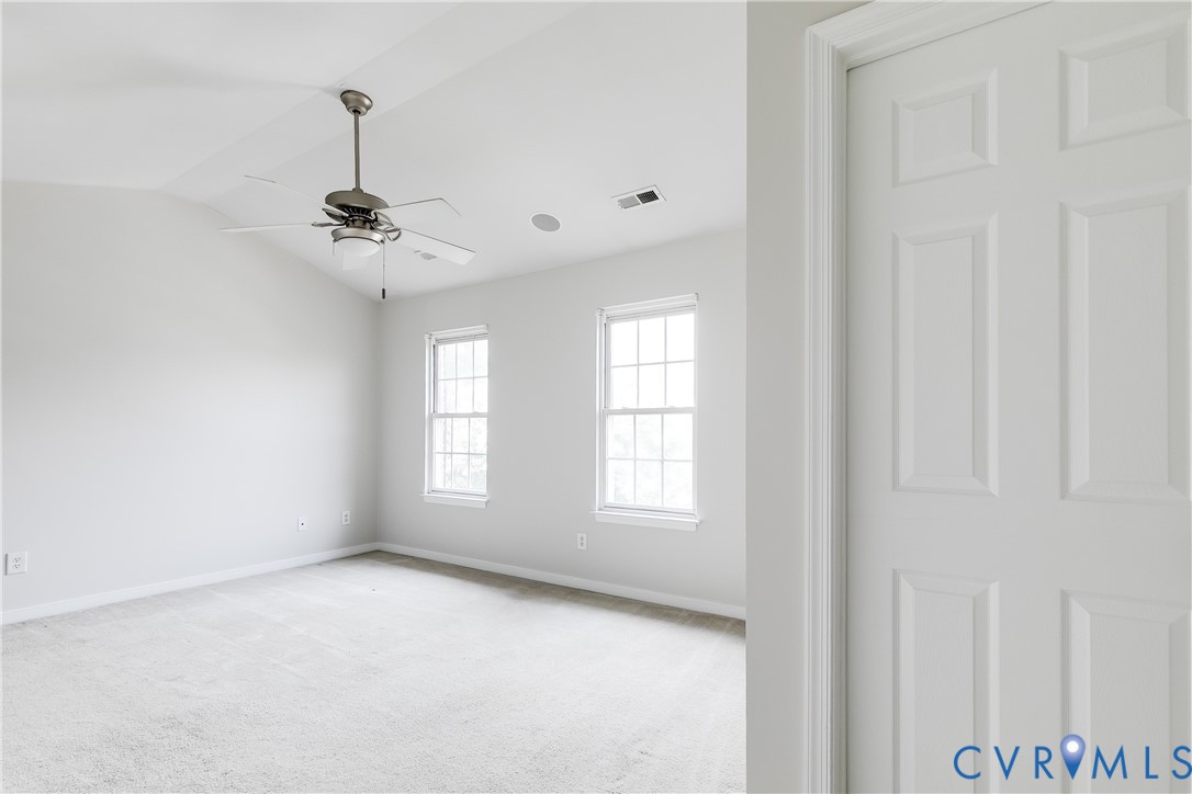 Spacious room featuring vaulted ceilings, a ceiling fan with integrated lighting, two double-hung windows, light-colored carpeting, and a six-panel interior door