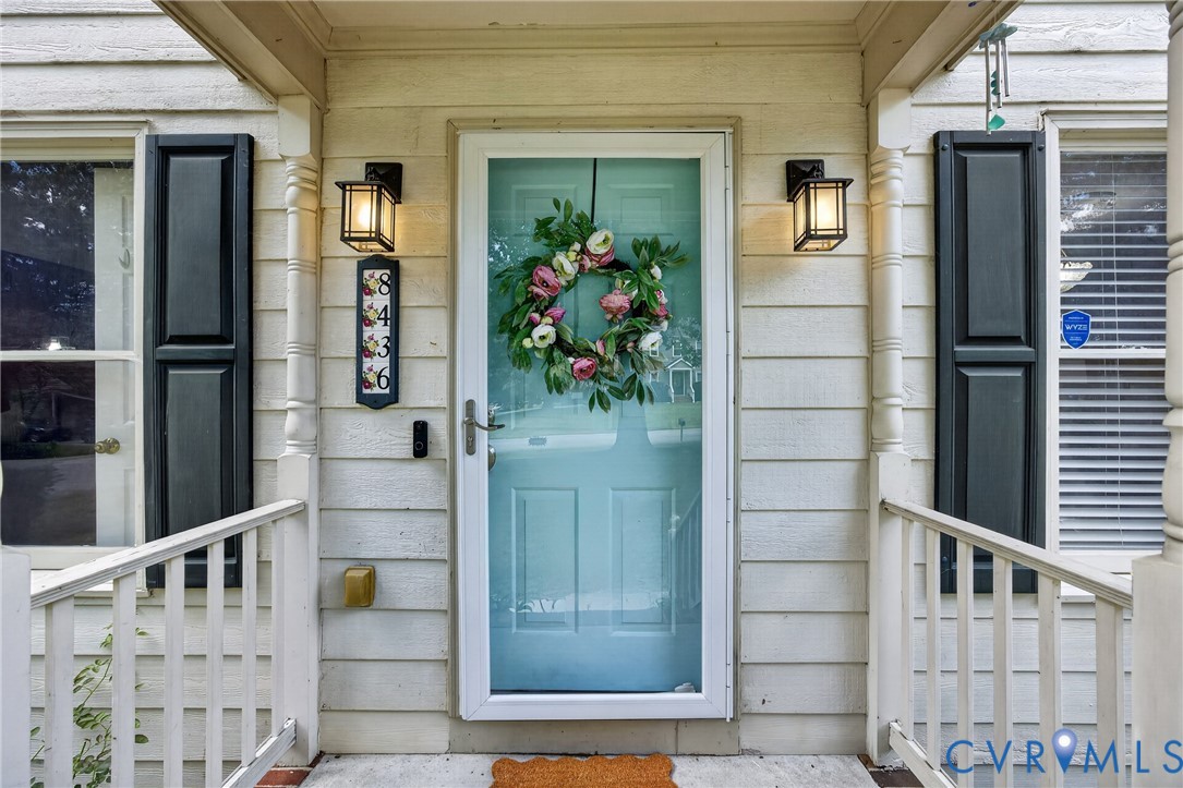 Light blue entry door with a white storm door, flanked by two windows with dark shutters, and a white porch railing