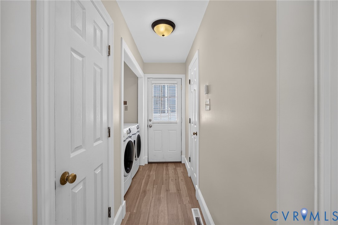 Bright laundry hallway featuring wood-finish flooring, white six-panel closet doors with brass hardware, a ceiling-mounted light fixture, and direct access to an exterior door with multiple storage closets lining the passage