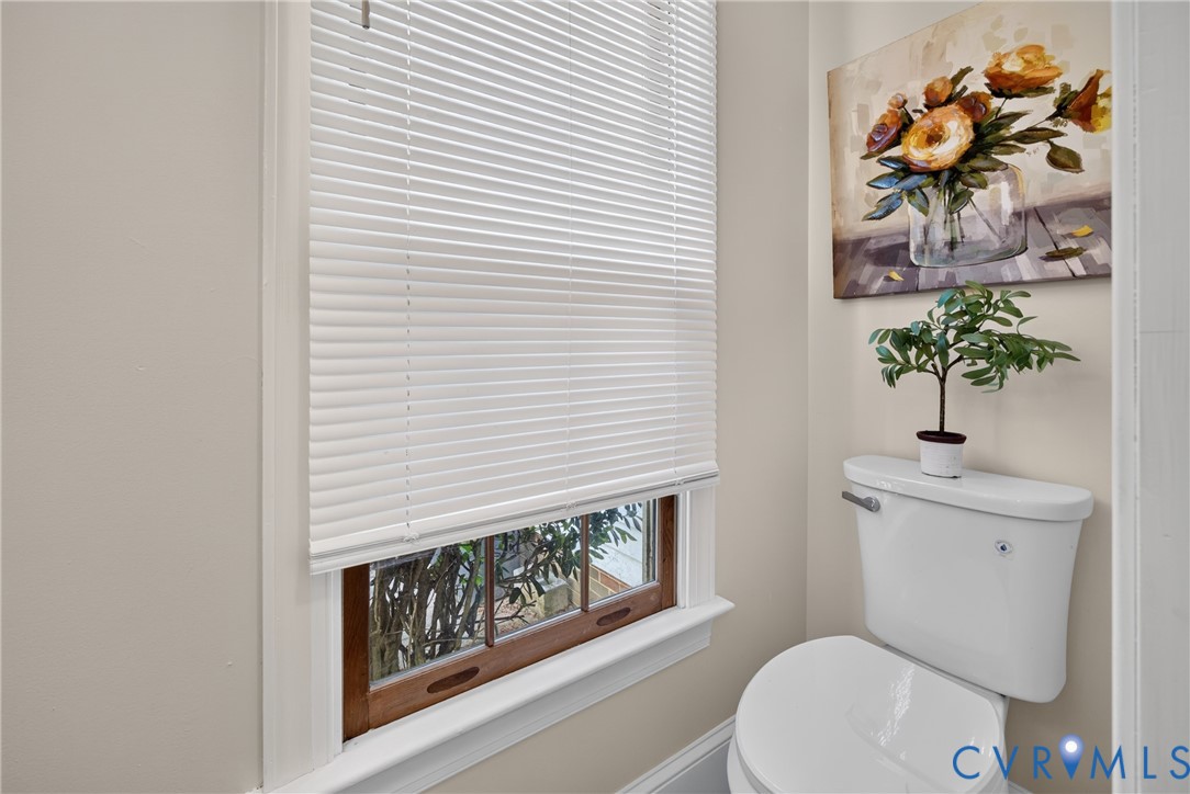 White toilet with a built-in flush handle, a window with wood-finish trim, and white horizontal blinds