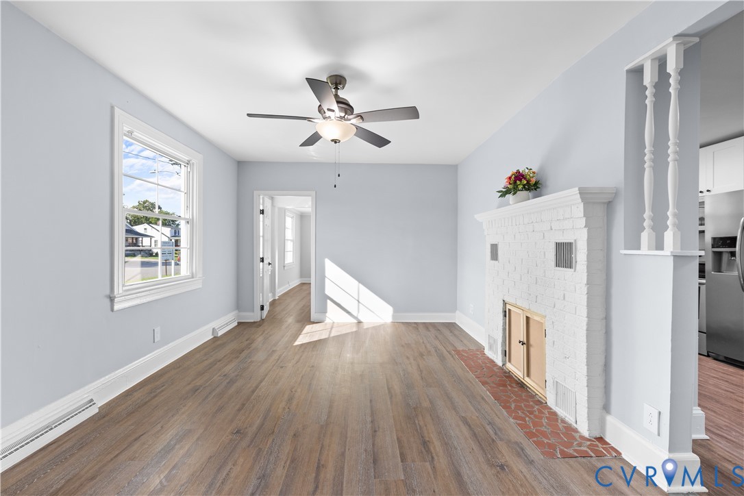 Wood-finish flooring, white painted brick fireplace with hearth, ceiling fan with light fixture, white trim, and a window with natural light