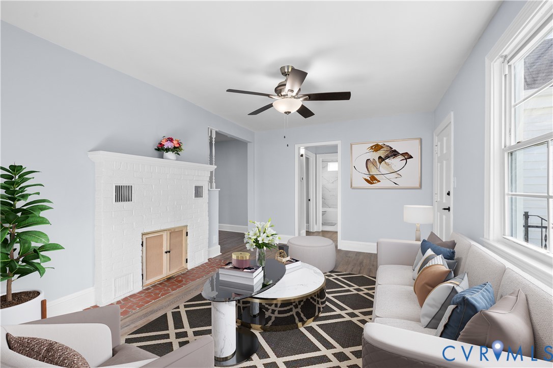 (Virtually-Staged)White painted brick fireplace, wood-finish flooring, ceiling fan with integrated lighting, white trim and window frames, and light-colored wall paint