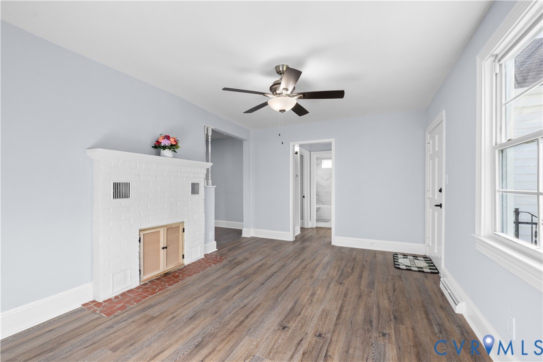 Wood-finish flooring throughout, white painted brick fireplace with brick hearth, ceiling fan with integrated lighting, paneled entry door, and double-hung window
