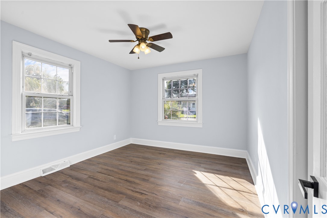 Light-filled room featuring wood-finish flooring, two large windows, a ceiling fan with integrated lighting, and a white door with matte black hardware