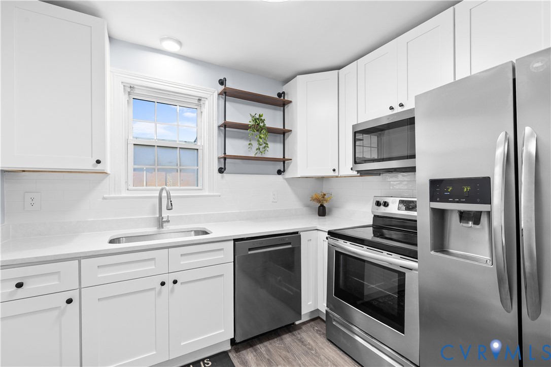 White shaker-style cabinetry, stainless steel appliances, light-toned countertops, subway tile backsplash, and wood-finish flooring White shaker-style cabinetry, stainless steel appliances, light-toned countertops, subway tile backsplash, and wood-finish flooring