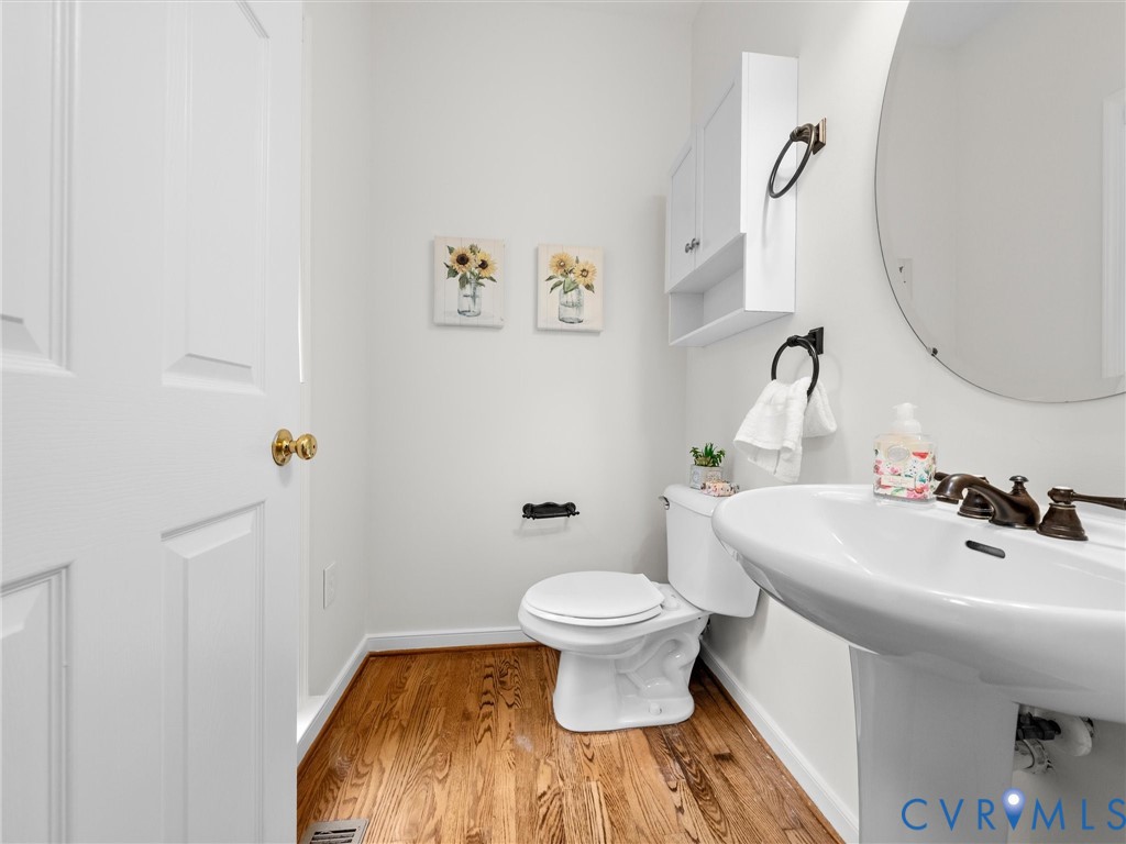 Wood-finish flooring, white pedestal sink, circular wall mirror, white toilet, and white wall-mounted cabinet