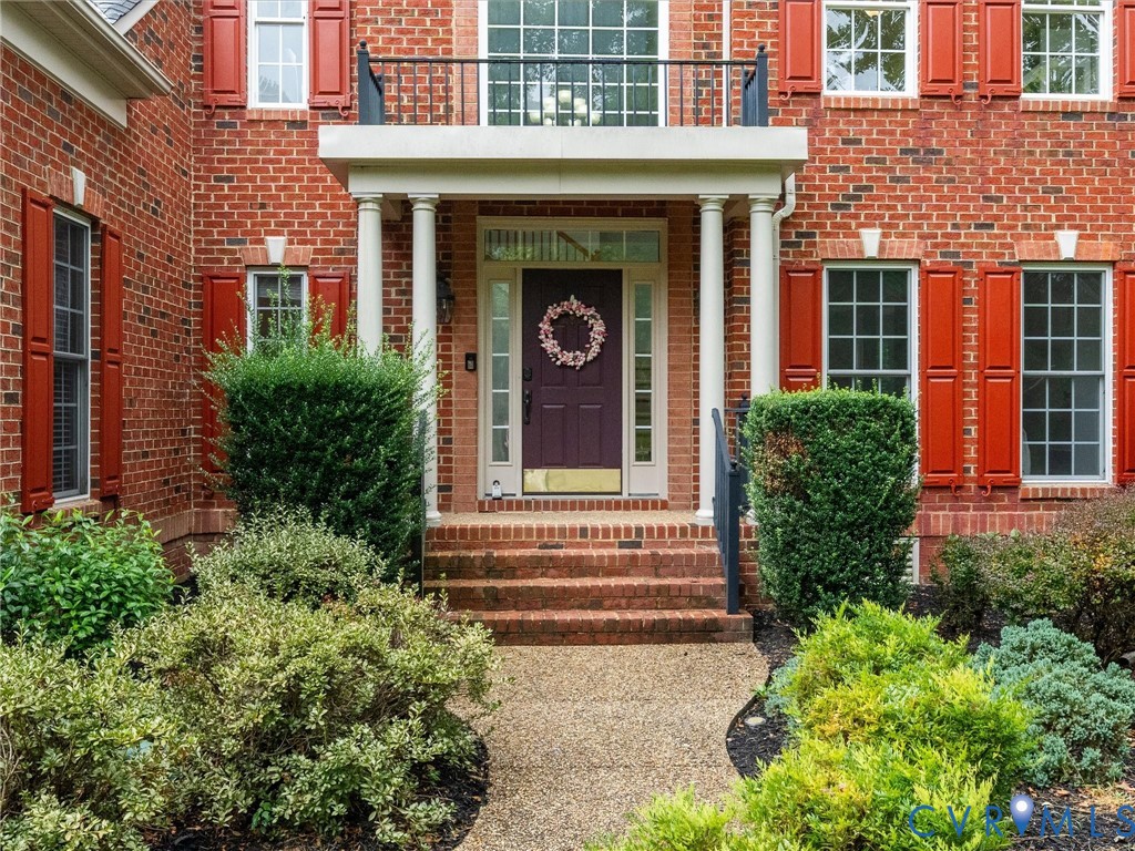 Brick facade with red shutters, portico with white columns, second-story balcony with black railing, and mature landscaping