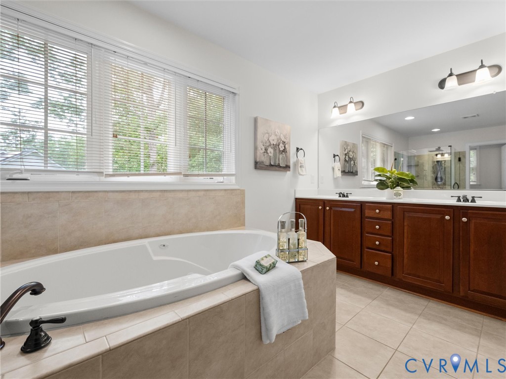 Built-in soaking tub with surrounding tile, dual vanity with wood cabinetry, and a large wall-mounted mirror