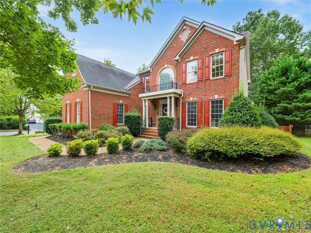 Brick exterior with red shutters, arched window, front porch with balcony, and mature landscaping