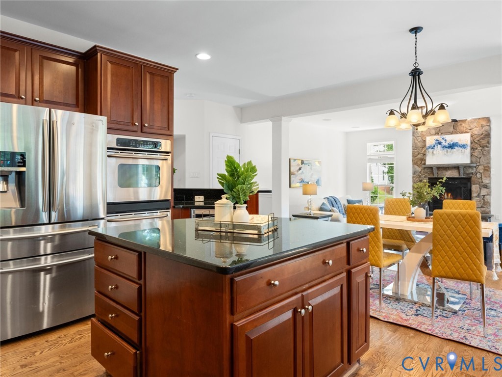 Hardwood-finish flooring, cherry wood cabinetry, stainless steel appliances, a kitchen island with a dark countertop, and a stone fireplace
