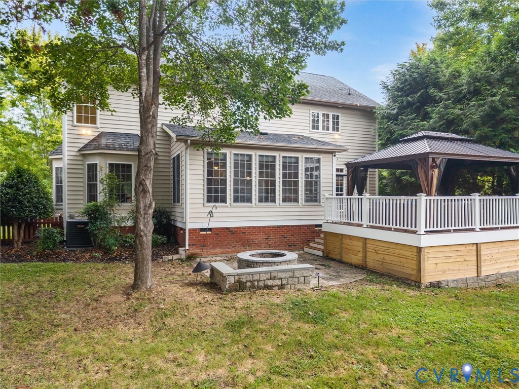 Rear exterior with siding, a sunroom with multiple windows, an elevated deck featuring white railings, a built-in gazebo, and a stone-lined fire pit on a paver patio